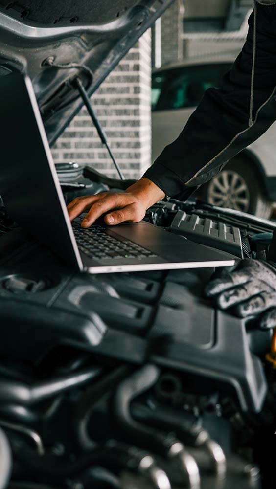 Auto shop service technician performing diagnostics with a technician using a laptop to scan a vehicle under the hood.
