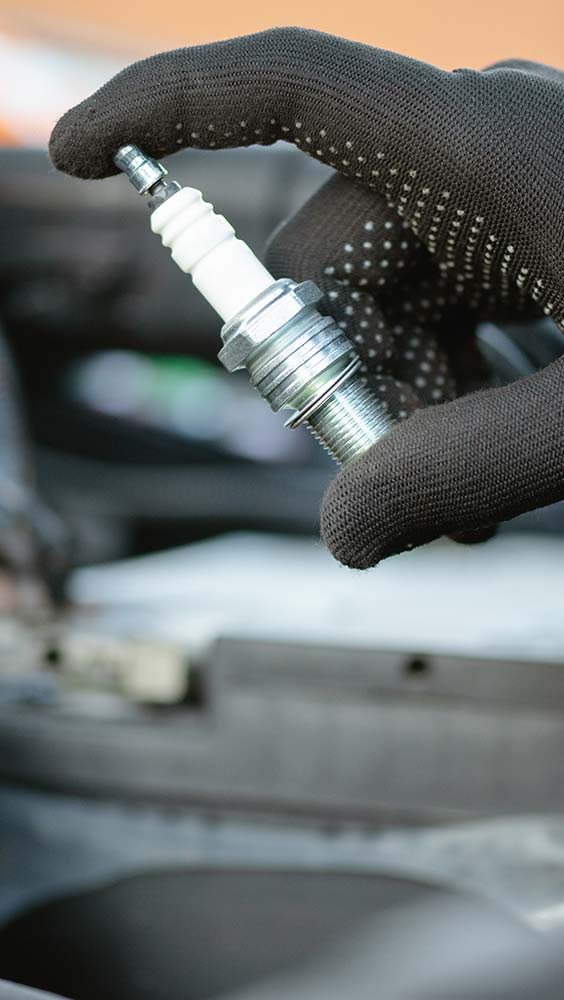 Ignition and fuel system repair showing a technician holding a spark plug near an engine bay.